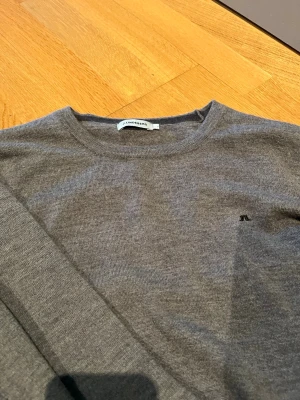 Grå stickad tröja från J.Lindeberg - Mjuk och stilren grå stickad tröja från J.Lindeberg med rund halsringning och diskret logga på bröstet. Tröjan har långa ärmar och ribbade muddar vid ärmslut och nederkant. Perfekt till lager på lager och enkel att matcha. Gjord av 100% ull