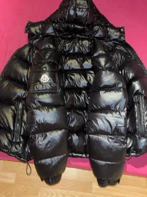 Svart glansig dunjacka från Moncler - Säljer en svart, glansig dunjacka från Moncler med stor huva och klassisk logotyp på bröstfickan. Jackan har quiltad design, dragkedjor på sidorna och justerbar nederkant. Perfekt för kalla vinterdagar och riktigt snygg streetstil.