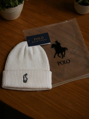 Vit ribbad mössa från Polo Ralph Lauren - 🧢 Vit ribbstickad beanie från Polo Ralph Lauren   ✨ Klassisk design med broderad logga 🐎   📦 Levereras i originalförpackning med tagg och plastpåse med logga   🌿 Mjuk, varm och stilren – perfekt för kalla dagar eller som fashion statement   🎁 Passar både dam & herr – one size  