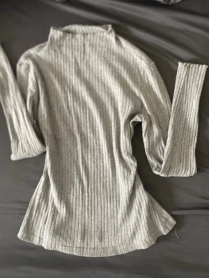 Ljusgrå ribbstickad polotröja - Säljer en ljusgrå ribbstickad polotröja med lång ärm och hög hals. Tröjan har en smal passform och är tillverkad i ett mjukt, stretchigt material som sitter snyggt på kroppen. Perfekt för lager-på-lager och enkel att matcha med olika outfits.