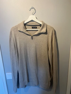 Beige halvzip tröja från Jack & Jones - Snygg beige stickad tröja från Jack & Jones med halvzip och hög krage. Tröjan har långa ärmar och ribbade muddar vid ärmslut och nederkant. Perfekt för lager på lager och enkel att matcha med olika outfits. Aldrig använd, säljes då den är för stor!
