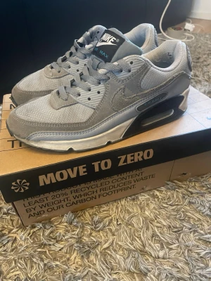 Nike Air Max 90 grå sneakers - Snygga Nike Air Max 90 sneakers i olika nyanser av grått. Ovandelen är i mesh och mocka med klassisk Nike Swoosh på sidan. Sulan har synlig Air Max-dämpning och detaljer i svart och vitt. Perfekta för dig som gillar streetwear och sportig stil.