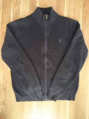Svart ribbstickad zip-kofta från Polo Ralph Lauren - Snygg och tidlös ribbstickad kofta med hel dragkedja från Polo Ralph Lauren. Klassisk svart färg med broderad logga på bröstet. Perfekt som lager på lager under höst och vinter eller som lättare jacka under svalare kvällar.  ✔️ Stilren och lättmatchad ✔️ Hel dragkedja ✔️ Broderad Polo-logga ✔️ Ribbad struktur för exklusiv känsla ✔️ Perfekt för både vardag och mer uppklätt