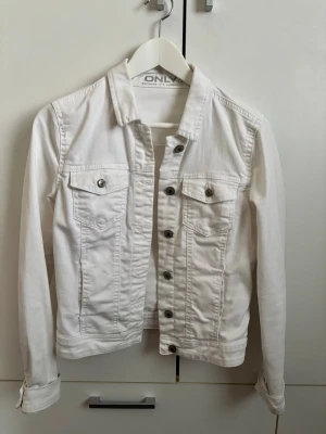 Vit jeansjacka från ONLY - Snygg vit jeansjacka från ONLY med klassisk krage, två bröstfickor med knappar och silverfärgade knappar framtill. Figurnära passform och långa ärmar. Perfekt att slänga över en t-shirt för en fräsch look.