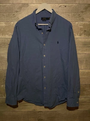Blå skjorta från Polo Ralph Lauren - Säljer en klassisk blå skjorta från Polo Ralph Lauren med button-down krage och broderad logga på bröstet. Skjortan har långa ärmar, knappar framtill och är tillverkad i bomull. Perfekt för dig som vill ha en stilren och tidlös look.
