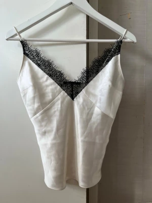 Vit satintopp med spets - Snyggt vitt satinlinne från Gina Tricot med tunna axelband och svart spetsdetalj längs v-ringningen. Toppen har en mjuk satinliknande yta och är perfekt för att skapa en chic och trendig look.