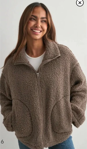 Brun teddyjacka  - Mysig brun teddyjacka med hög krage och hel dragkedja framtill. Jackan har två stora fickor och är tillverkad i fluffigt, mjukt material som ger en avslappnad och trendig look. Perfekt för att hålla sig varm under kyliga dagar.