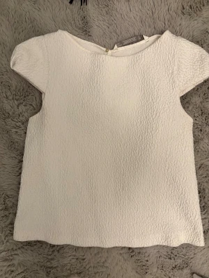 Vit strukturerad blus från Zara - Säljer en vit blus från Zara med korta ärmar och en snygg strukturerad yta. Toppen har rund halsringning och knappdetalj i nacken. Perfekt till jeans eller kjol för en clean look.
