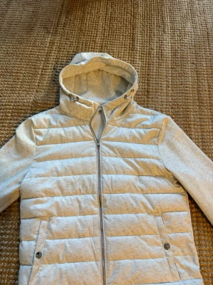 Ljusgrå quiltad vindjacka med huva - Snygg ljusgrå vindjacka med quiltat mönster på kroppen och mjuka ärmar. Jackan har dragkedja framtill, två fickor med tryckknappar och en justerbar huva med snörning. Perfekt för blåsiga dagar och enkel att matcha med olika outfits.