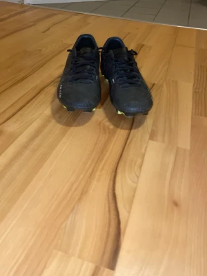 Svarta Nike fotbollsskor med dobbar - Säljer ett par svarta Nike fotbollsskor med gula dobbar och detaljer. Skorna har snörning och är tillverkade i syntetmaterial med en sportig design och Nike-logga på sidan. Perfekta för spel på gräsplaner och ger bra grepp och kontroll.
