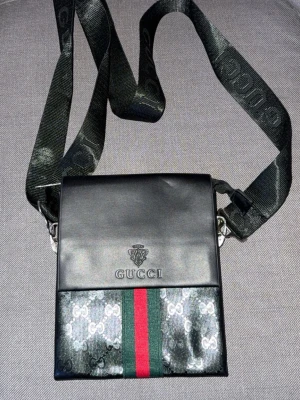 Svart axelväska från Gucci - Snygg svart axelväska från Gucci med klassiskt GG-mönster och gröna samt röda ränder framtill. Hitta den i min garderob då jag är osäker på pris säljer jag billigt. Väskan har justerbar axelrem med logga och dragkedjeficka på baksidan. Tillverkad i slitstarkt material med stilren design, perfekt för att bära det viktigaste. På baksidan finns det vita fläckar då jag inte testat ta bort och inte vet vad det är sänker jag priset för det också.
