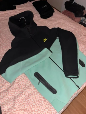 Nike tech  - Snygg hoodie från Nike i svart och mintgrönt med dragkedja framtill och huva. Har två fickor med dragkedja och en gul Nike-logga på bröstet. Perfekt för dig som gillar sportig och avslappnad stil.