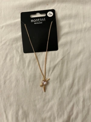 Guldigt halsband med hjärthänge - Snyggt guldigt halsband från Ur och Penn  med ett hänge i form av ett hjärta med ljusrosa sten och små detaljer som pilar och stavar. Kedjan är tunn och elegant, perfekt för att ge din outfit en söt touch. Materialet är metall med glittrande sten i hänget.