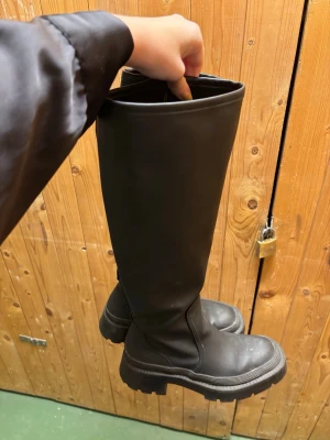 Zara höga svarta stövlar - Säljer ett par svarta höga stövlar med chunky sula och rund tå. Stövlarna är i ett slätt material som liknar skinn eller syntet och har en stilren, minimalistisk design utan synliga detaljer eller dekorationer. Perfekta för en edgy och trendig look.