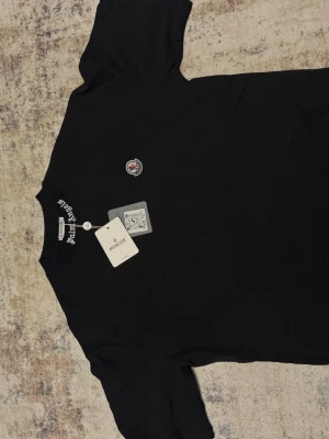 Svart Moncler x Palm Angels t-shirt - Svart t-shirt från Moncler i samarbete med Palm Angels. Stor vit text 'MONCLER 1952' och Palm Angels-logga på ryggen. Liten Moncler-patch på bröstet. Klassisk passform och rund hals. Tillverkad i mjuk bomull, perfekt för en clean och trendig look.