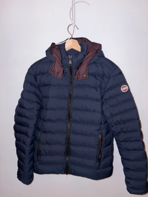 Mörkblå pufferjacka från Colmar - Snygg mörkblå pufferjacka från Colmar med bruna detaljer vid huvan och dragkedjor. Jackan har en vadderad design, två fickor med dragkedja och Colmar-logga på ärmen. Perfekt för kalla dagar och riktigt trendig stil.