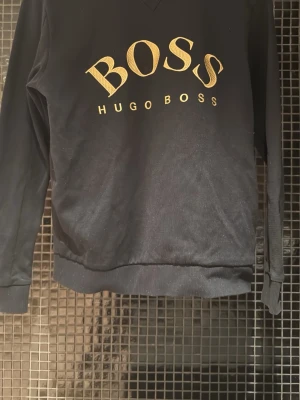 Svart sweatshirt från Hugo Boss - Cool svart sweatshirt från Hugo Boss med stort guldigt BOSS-tryck på bröstet och ribbade muddar vid ärmslut och nederkant. Perfekt för dig som gillar stilrena och trendiga plagg med tydlig logga.