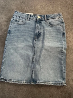 Ljusblå jeanskjol med fickor - Säljer en klassisk ljusblå jeanskjol med fem fickor och knappstängning fram. Kjolen har en rak passform och är tillverkad i denim med snygga sömdetaljer. Perfekt att styla med sneakers eller boots. Köptes för 349 kr