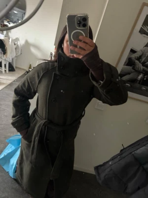 Mörkgrön ullkappa med hög krage - Snygg mörkgrön ullkappa med dubbelknäppning och hög krage. Kappan har ett knytskärp i midjan och långa ärmar med dekorativa detaljer vid ärmsluten. Perfekt för kalla dagar och ger en stilren siluett. Köpt för 900. Mörkgrön färg