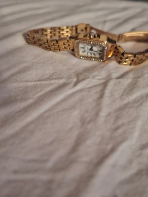 Guldfärgad fyrkantig klocka - Stilren guldfärgad klocka med fyrkantig urtavla och romerska siffror. Klockan har en länkad metallrem och detaljer runt urtavlan som ger en lyxig känsla. Perfekt accessoar för att lyfta din outfit.