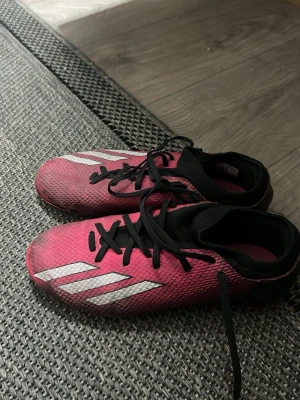 Rosa Adidas fotbollsskor med svarta detaljer - Snygga fotbollsskor från Adidas i en cool rosa färg med svarta snören och detaljer. Skorna har de klassiska vita Adidas-ränderna på sidan och en ovandel i slitstarkt syntetmaterial. Perfekta för dig som vill sticka ut på planen!