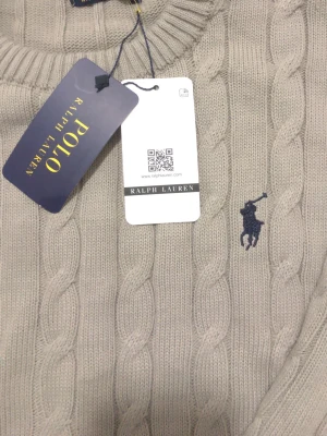Beige kabelstickad tröja Polo Ralph Lauren - Kabelstickad beige tröja från Polo Ralph Lauren med klassisk rund halsringning och broderad marinblå logga på bröstet. Tröjan har långa ärmar och ribbade muddar vid ärmslut och nederkant. Perfekt för en stilren och avslappnad look.