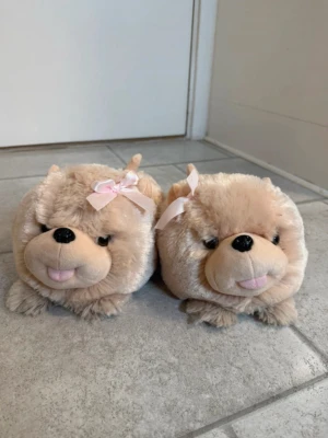 Beige fluffiga hundtofflor med rosett - Supersöta beige tofflor i plyschmaterial formade som hundar med små öron, nos och tunga. Båda tofflorna har en ljusrosa rosett på huvudet för extra gullig look. Perfekta för att hålla fötterna varma hemma.