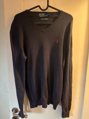 Mörkblå v-ringad tröja Polo Ralph Lauren - Mörkblå v-ringad tröja från Polo Ralph Lauren med klassisk röd broderad logga på bröstet. Tröjan har långa ärmar och ribbade muddar vid ärmslut och nederkant. Perfekt för dig som gillar stilrena och tidlösa plagg.