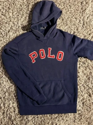 Marinblå hoodie från Polo Ralph Lauren🤩🙌 - Snygg marinblå hoodie från Polo Ralph Lauren med stor röd POLO-text på bröstet. Klassisk känguruficka framtill och huva. Tillverkad i mjukt bomullsmaterial som är perfekt för chill dagar. Passar dig som gillar stilrena och sportiga plagg.🙌