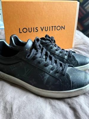 Svarta Louis Vuitton sneakers - Model Luxumborg, Finns låda till. Snygga svarta sneakers från Louis Vuitton med klassiskt monogrammönster i grått på lädret. Skorna har svarta skosnören, vit sula och diskreta LV-detaljer på sidan. Perfekta för dig som vill ha en exklusiv och stilren look. LV size 7