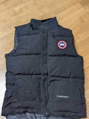 Svart dunväst från Canada Goose - Snygg svart dunväst från Canada Goose med klassisk logotyp på bröstet och två stora fickor framtill. Västen har hög krage och stängs med tryckknappar. Perfekt för lager-på-lager och håller dig varm under kyliga dagar.