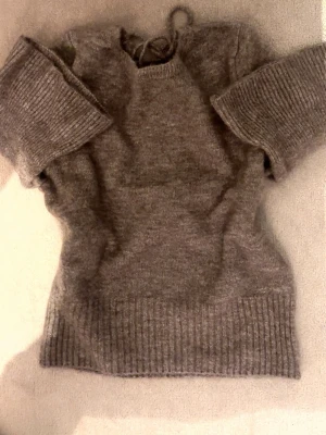 Grå/brun stickad tröja med öppen rygg - Mysig grå/brun stickad tröja med korta ärmar och bred ribbad kant nedtill. Tröjan har en öppen rygg med knytband upptill. Perfekt för lager på lager och passar till både jeans och kjol.