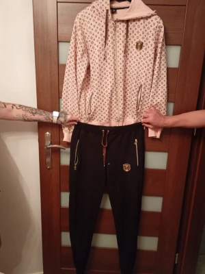 Beige och svart LV loungewear set - Säljer ett snyggt loungewear set från Louis Vuitton med beige hoodie med monogrammönster och dragkedja samt svarta byxor med gulddetaljer och dragsko i midjan. Båda delarna har LV-logga och mjukt material som passar perfekt för chill dagar.