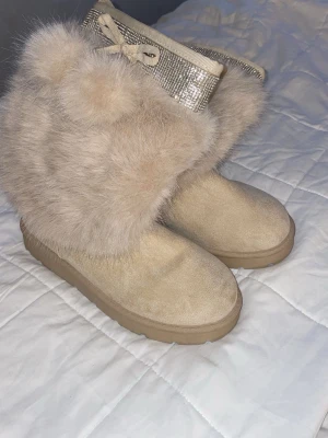 Beige stövletter med fuskpäls och glitter - Säljer ett par beige stövletter med fluffig fuskpäls och glittrigt skaft med snörning upptill. Skorna har en robust sula och är tillverkade i syntetmaterial och textil. Perfekta för dig som vill ha något extra och mysigt på fötterna.