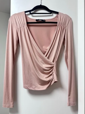Ljusrosa omlottopp från Bik Bok - Supersnygg ljusrosa omlottopp från Bik Bok med lång ärm och v-ringning. Toppen har rynkade detaljer i sidan och axlarna, vilket ger en cool och trendig look. Perfekt att styla med jeans eller kjol för en chic outfit.