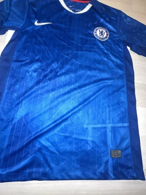 Chelsea FC blå matchtröja från Nike stl m - Chelsea FCs officiella blå matchtröja från Nike med klubbmärke på bröstet och vit Nike-logga. Tröjan har tunna vertikala ränder och är tillverkad i ett lätt, ventilerande material som passar perfekt för fotbollsträning eller match.