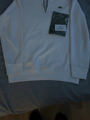 Vit half-zip hoodie från Lacoste - Snygg vit hoodie från Lacoste med half-zip dragkedja och klassiska krokodilloggan broderad på bröstet. Hoodien har en stor magficka och ribbade muddar vid ärmar och nederkant. Tillverkad i mjukt material som känns skönt mot huden.