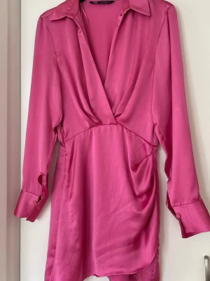 Rosa klänning från Zara - Snygg rosa kortklänning från Zara i omlottmodell med v-ringning och långa ärmar. Tillverkad i ett glansigt material som ger en lyxig känsla. Perfekt för dig som vill sticka ut med en färgstark look.