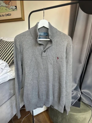 Grå stickad tröja Polo Ralph Lauren - Snygg grå stickad tröja från Polo Ralph Lauren med ribbad krage och två knappar vid halsen. Tröjan har lång ärm och den klassiska röda logotypen broderad på bröstet. Tillverkad i 100% bomull för en mjuk och bekväm känsla.