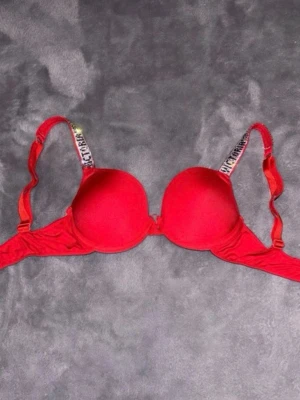 Röd push-up bh från Victoria's Secret - Snygg röd push-up bh från Victoria's Secret med vadderade kupor och justerbara axelband med logotyp. Bh:n har en slät design och ger bra stöd. Perfekt för dig som vill ha en bekväm och trendig bh med lite extra lyft. Se sista bilden för att se defekter. Defekten påverkar inte användningen. Tror det är C kupa men skulle säga 70C