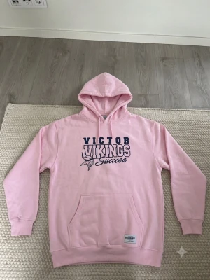 Rosa hoodie Victor Vikings Gildan - Snygg rosa hoodie från Gildan med trycket 'Victor Vikings Success' i blått och vitt på bröstet. Klassisk känguruficka framtill och justerbar huva. Tillverkad i mjuk bomullsblandning, perfekt för chill dagar eller när du vill sticka ut lite extra.