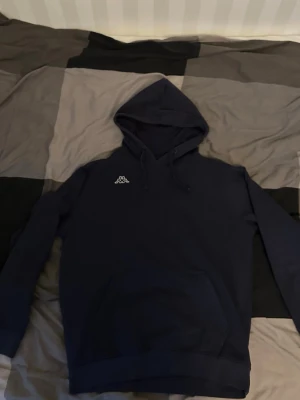 Mörkblå hoodie från Kappa - Snygg mörkblå hoodie från Kappa med klassisk logga broderad på bröstet. Hoodien har huva med dragsko och en stor magficka framtill. Perfekt för en chill och avslappnad stil. Tillverkad i mjukt material för extra komfort.