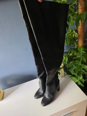 Svarta overknee stövlar med klack - Säljer ett par svarta overknee stövlar i slätt konstläder. De har rundad tå och bred klack som ger stabilitet och höjd. Stövlarna är höga och går över knät, perfekta för en trendig look till hösten eller vintern.