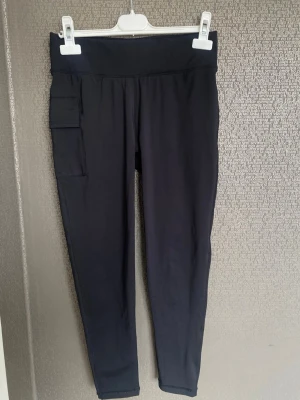 Svarta leggings med ficka - Snygga svarta leggings med hög midja och praktisk sidoficka på vänster ben. Tillverkade i stretchigt material som sitter skönt och följer kroppen. Reflekterande detaljer baktill vid vaderna för extra stil och synlighet.