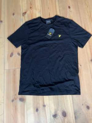 Svart t-shirt från lyle skott - Svart t-shirt med rund hals och korta ärmar. På bröstet finns en liten gul broderad logga. Enkel och stilren design som passar till det mesta. Tillverkad i mjukt material för skön känsla.