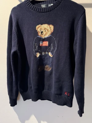 Polo Ralph Lauren tröja Bear - Mörkblå stickad tröja från Ralph Lauren med den ikoniska Polo Bear på bröstet, bärande en tröja med amerikanska flaggan. Rund halsringning och ribbade muddar. RL-broderi vid nederkanten. Passar till det mesta, båda uppklätt och lugnare aktiviteter. Strl S, 67% bomull, 33% linne. Skickas snabbt och säkert via Plick, hör av er vid frågor