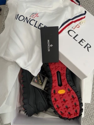 Moncler Grenoble svarta sneakers med röd sula - Svarta sneakers från Moncler med robust design och grov röd Vibram-sula. Skorna har detaljer i Gore-Tex för extra funktionalitet och är perfekta för dig som vill ha både stil och prestanda. Levereras med originalpåse och box.