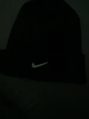 Svart mössa från Nike - Säljer en svart ribbstickad mössa från Nike med klassisk vit swoosh-logga framtill. Mössan har uppvikt kant och är tillverkad i mjukt material, perfekt för kalla dagar. Enkel och stilren design som passar till det mesta.