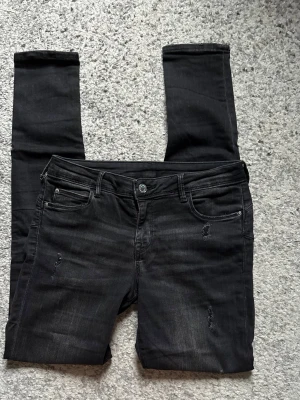 Svarta skinny jeans med slitningar - Säljer ett par svarta skinny jeans med diskreta slitningar framtill. Jeansen har klassisk femficksmodell, knappgylf och smal passform. Materialet är stretchigt för extra komfort och sitter tight hela vägen ner.