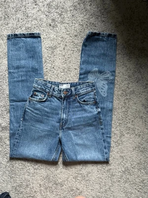 Blå raka jeans med fjärilstryck - Snygga blå jeans med rak passform och hög midja. Jeansen har klassisk femficksdesign och ett unikt vitt fjärilstryck på ena benet. Tillverkade i denim med en cool vintagekänsla. Perfekta för dig som vill sticka ut med detaljer.